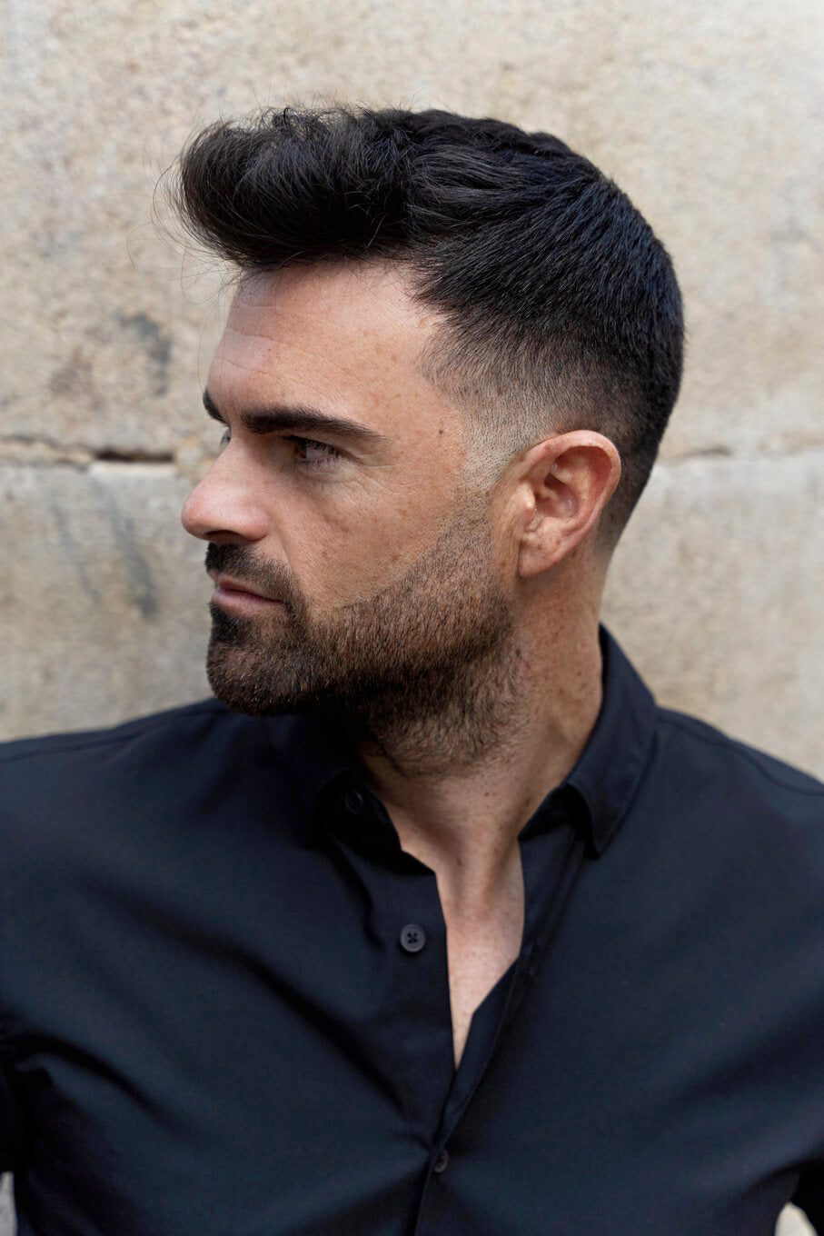 Hombre con cabello corto degradado en los laterales y peinado voluminoso en la parte superior, luciendo un estilo contemporáneo con camisa negra.